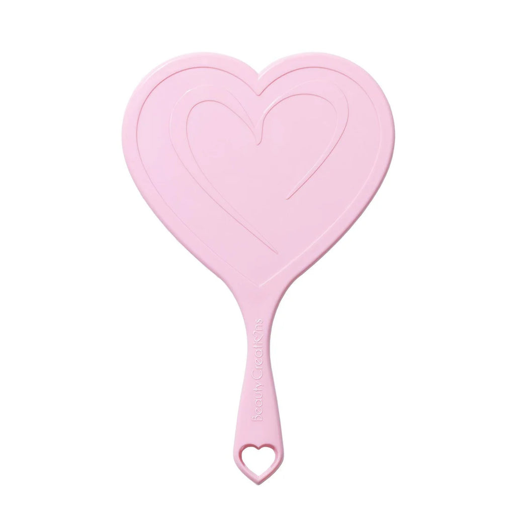 Un espejo de mano en forma de corazón de color rosa claro con mango que presenta un pequeño recorte de corazón en el extremo, mostrado sobre un fondo blanco limpio.