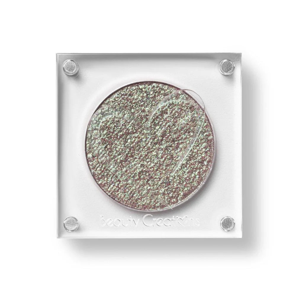 Un estuche cuadrado de sombra de ojos de Beauty Creations en acrílico transparente con un hueco redondo de sombra plateada con destellos verdes y rosa brillante, con un recorte en forma de corazón en la tapa.