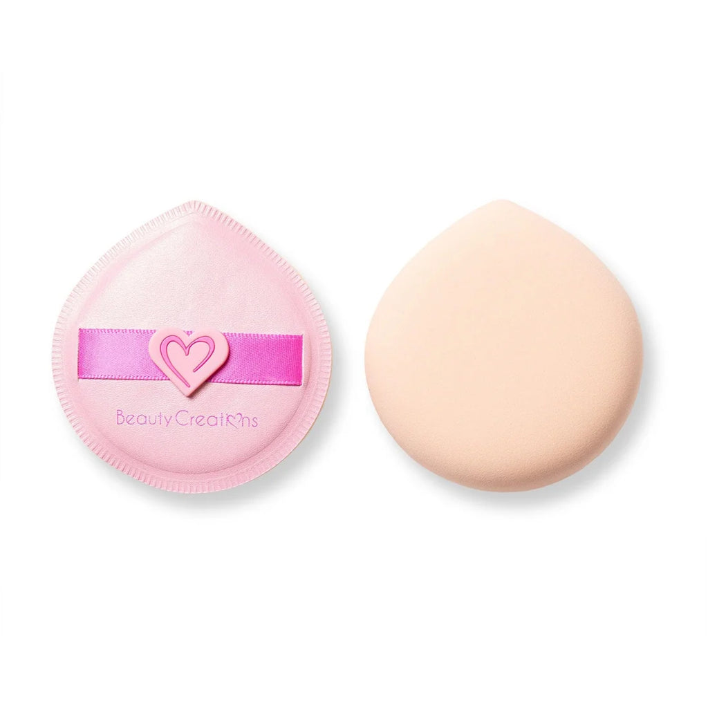Dos esponjas redondas de polvo para maquillaje sobre un fondo blanco: una esponja rosa de Beauty Creations con una correa en forma de corazón junto a una esponja melocotón lisa y de aspecto suave.