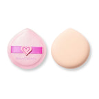 Dos esponjas redondas de polvo para maquillaje sobre un fondo blanco: una esponja rosa de Beauty Creations con una correa en forma de corazón junto a una esponja melocotón lisa y de aspecto suave.