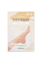Un empaque crema de las mascarillas para pies Shea-Socks de Shea Butter muestra un pie descalzo entre suaves formas florales, sugiriendo un cuidado de la piel relajante, tipo spa.