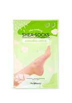 Un empaque verde de las mascarillas para pies Shea-Socks de Cannabis Sativa muestra un pie descalzo entre suaves formas florales, sugiriendo un cuidado de la piel relajante, tipo spa.